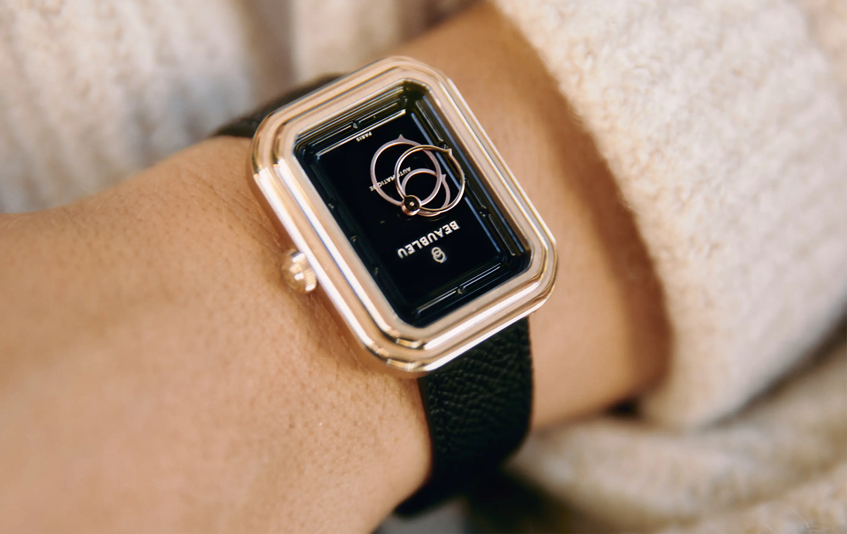 Montre automatique Figura Onyx en acier or rose avec bracelet cuir noir, portée au poignet d’une femme avec un pull beige