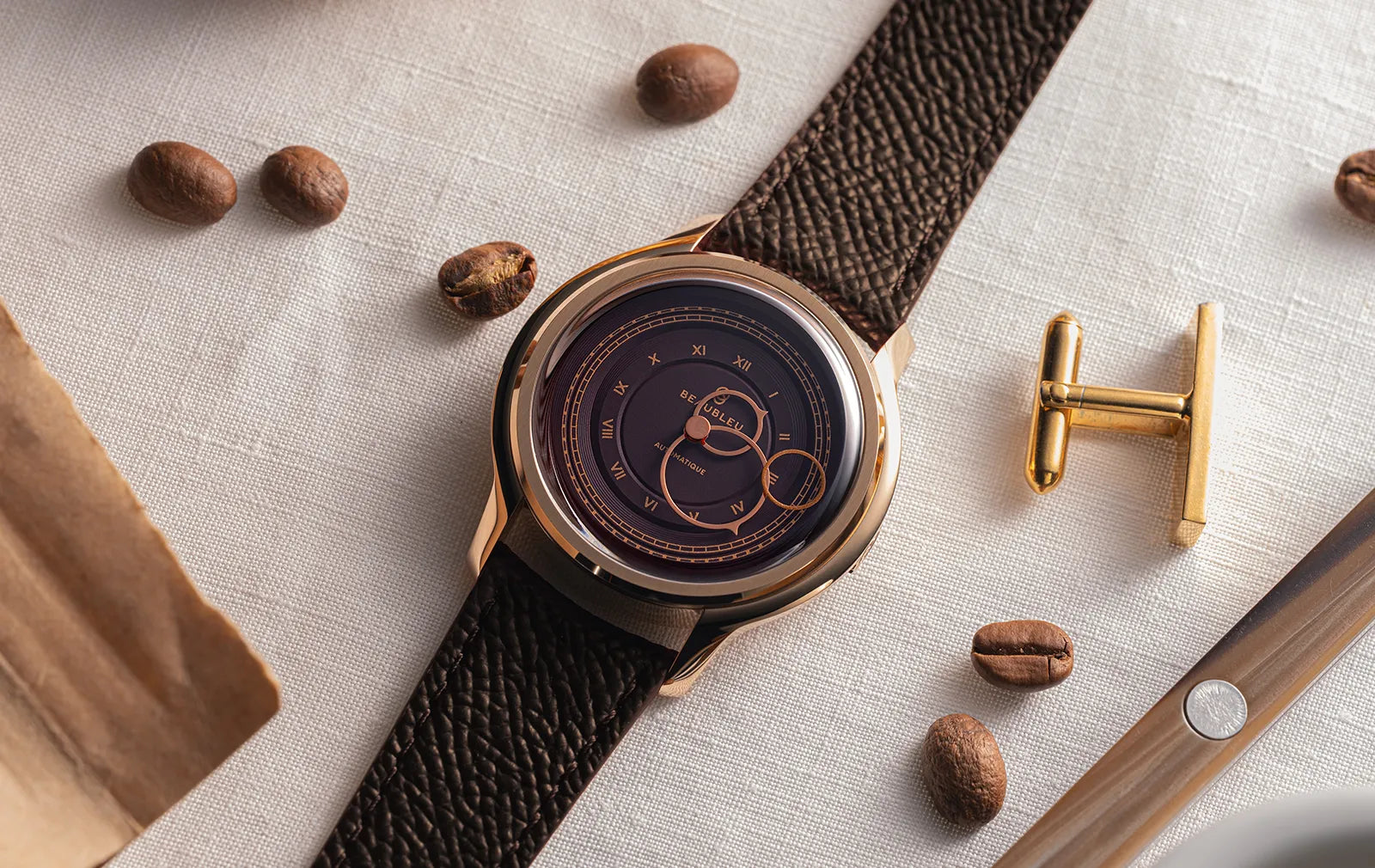 Montre dorée à cadran marron sur bracelet en cuir marron, entourée de grains de café et de boutons de manchette dorés