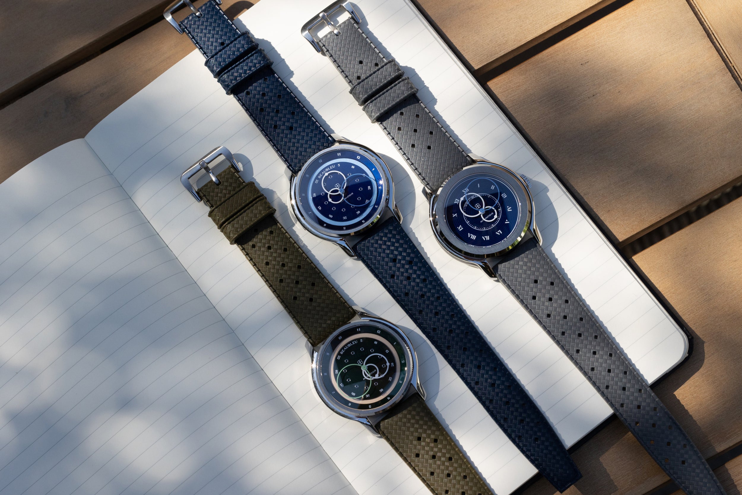 Trois montres Beaubleu : GMT Verte, GMT Bleue et Origine Bleue, avec bracelet tropic, posées sur un carnet.