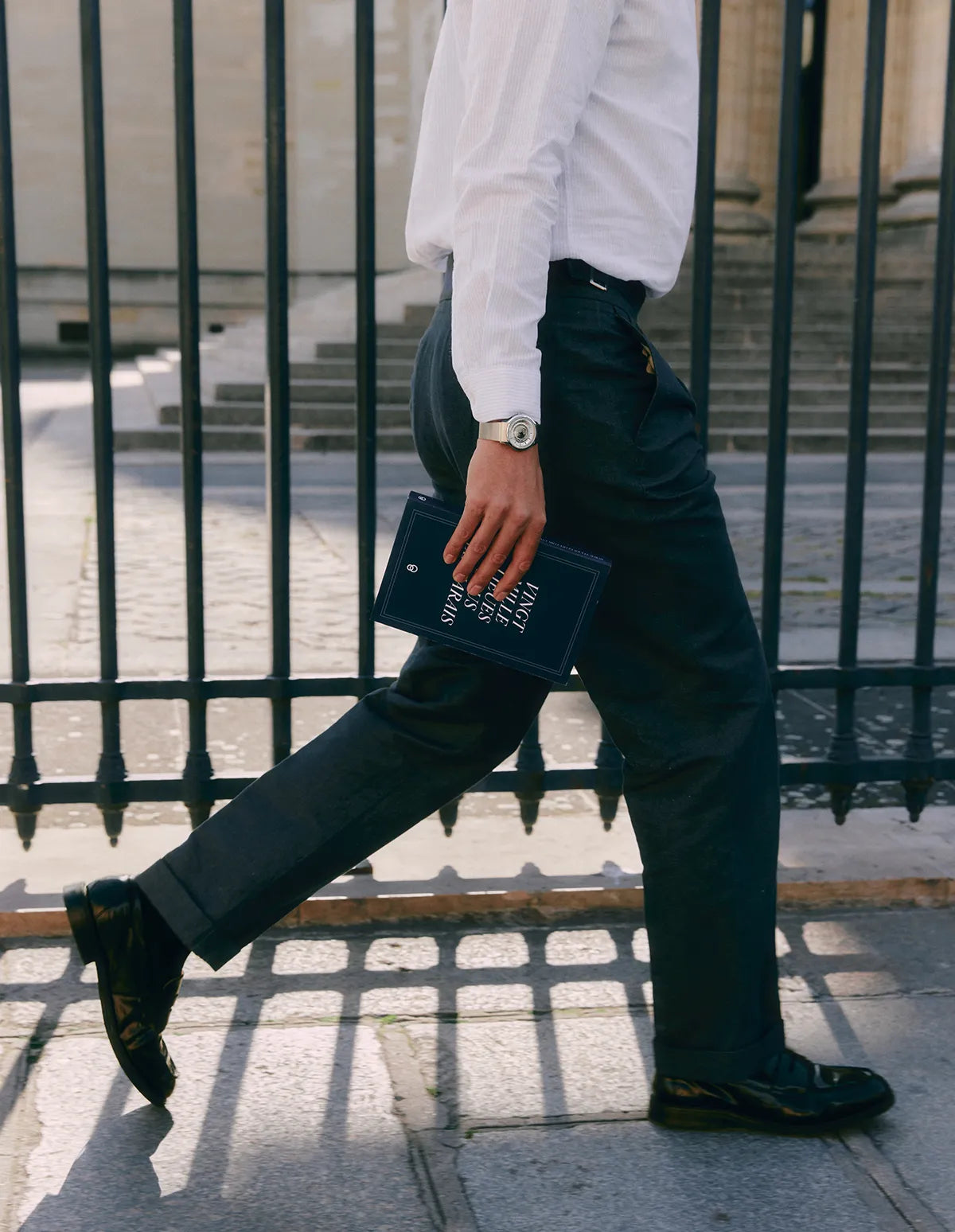 Montre Beaubleu La Pièce Acier portée au poignet d'un homme en costume marchant dans la rue avec un livre à la main